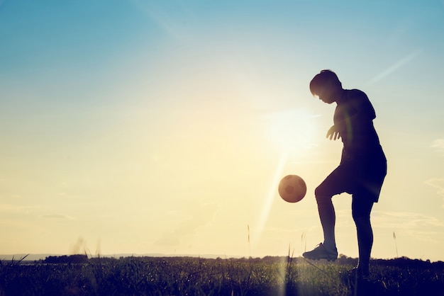 夕日の背景にサッカーボールを持つサッカー選手のシルエット。