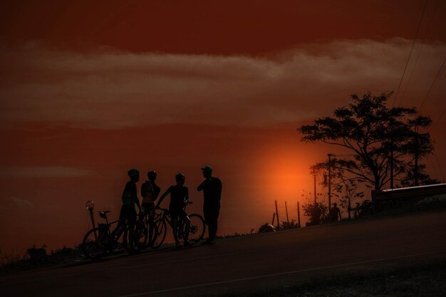 Silhouette people by bicycle on road against sky during sunset