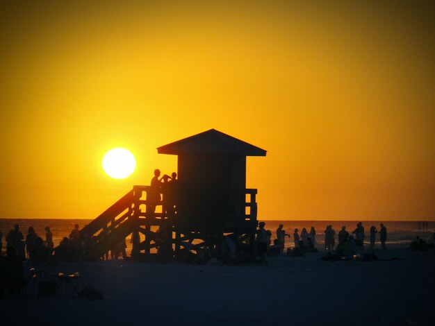 Silhouette people and lifeguard tower at beach against clear sky during sunset