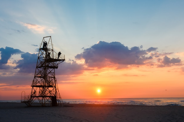 Silhouette of people on a rescue tower on a sandy beach at sunset.