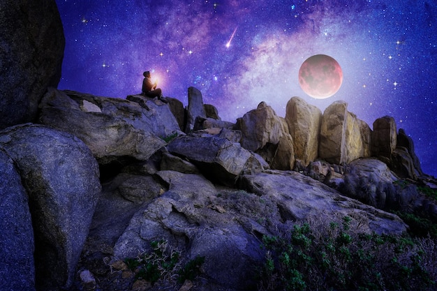 Silhouette of a person meditating or praying outdoors on a\
stone at night with stars and milky way