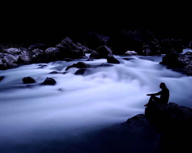Photo silhouette person sitting on rock by waterfall