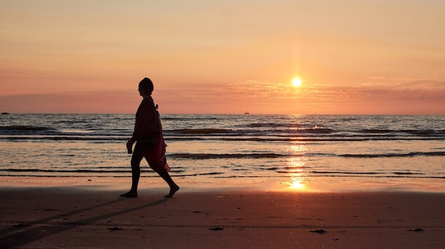 A silhouette of a person walking on a sea shore at sunset