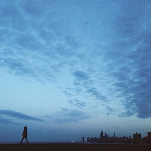 silhouette person walking on shore with cityscape in background against sky