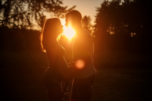 妊娠中の若い女性と夕暮れ時の屋外歩行の夫のシルエット。夏の夜に自然に手を取り合って幸せな家族。出産を待っている妊娠中の親と晴れた日にお互いを楽しむ