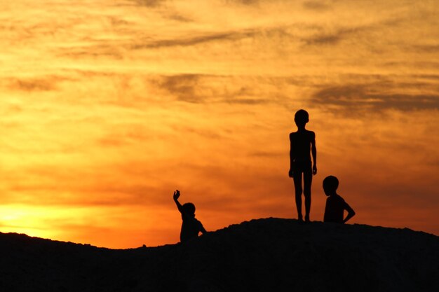 Silhouette siblings standing on hill against cloudy sky during sunset