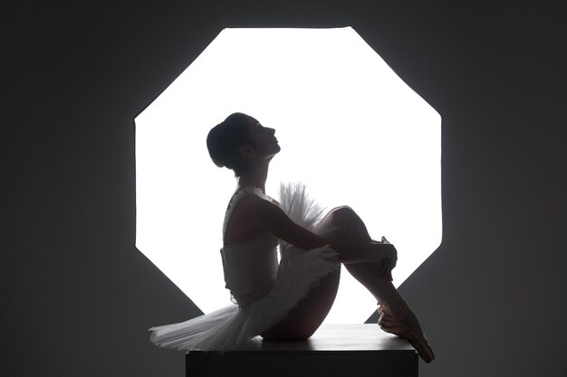 Silhouette of slim ballerina in tutu sitting on a cube against octabox in studio