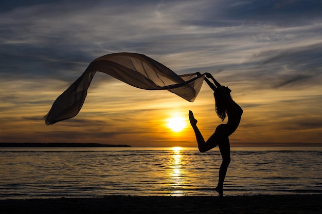 Silhouette of slim sexy girl dancing with scarf on the beach at sunset