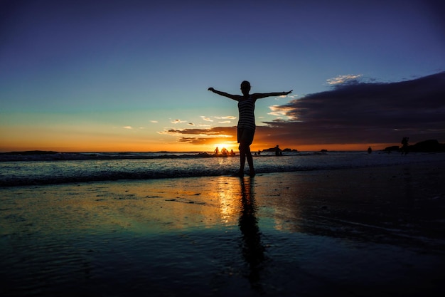 Силуэт стройной женщины с поднятыми руками, смотрящей на закат над морем на пляже