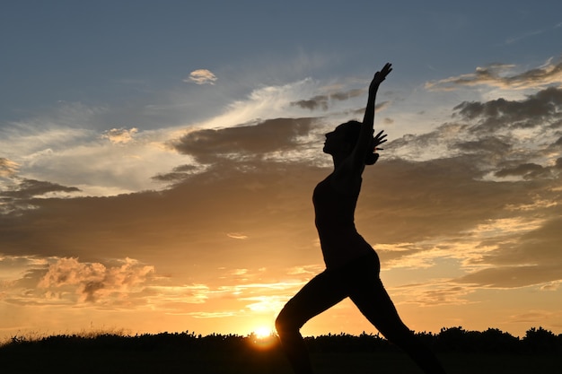 Силуэт стройной молодой женщины, практикующей позу воина йоги, закат на заднем плане