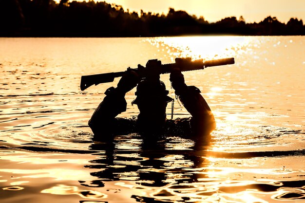 Силуэт спецназа с винтовкой в действии во время рейда, пересекающего реку в джунглях по пояс в воде, с оружием над головой