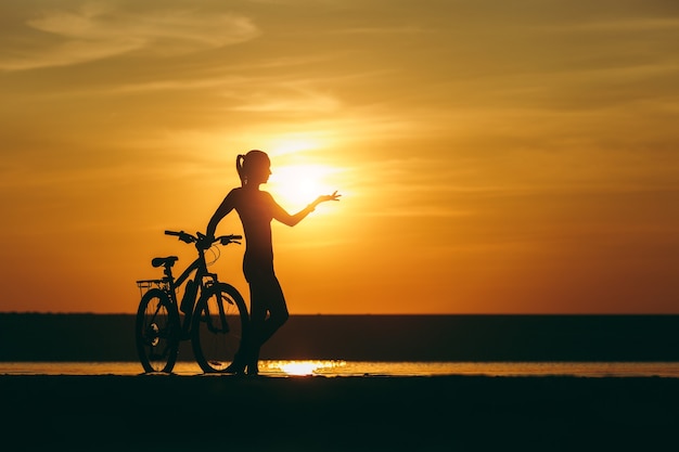 La silhouette di una ragazza sportiva in un vestito in piedi vicino a una bicicletta nell'acqua e punta la mano in lontananza al tramonto in una calda giornata estiva. concetto di forma fisica.