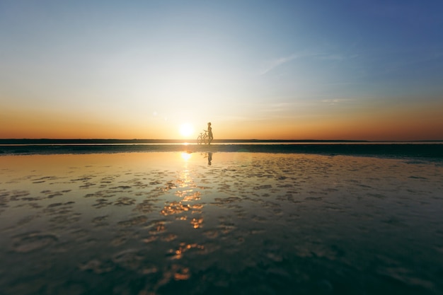 La silhouette di una ragazza sportiva in un vestito in piedi vicino a una bicicletta in acqua al tramonto in una calda giornata estiva. concetto di forma fisica.