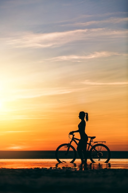 The silhouette of a sporty girl in a suit that sits on a bicycle in the water at sunset on a warm summer day. Fitness concept.