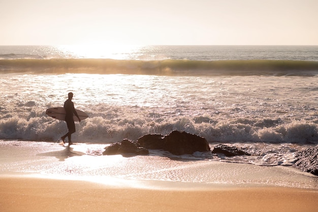 Silhouette of a surfer walking along the shore of the beach with his board under his arm at sunset concept of leisure and relax copy space for text