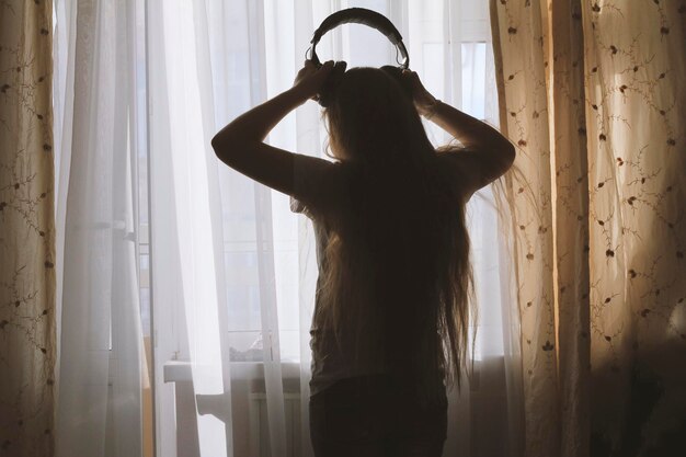 Silhouette of teenager girl wearing headphones in front of the window, concept of youth leisure