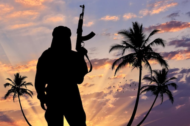 Silhouette of a terrorist with a weapon against a background of a sunset with palm trees