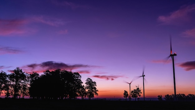 The silhouette of three modern windmills in a colorful sunset next to a group of trees