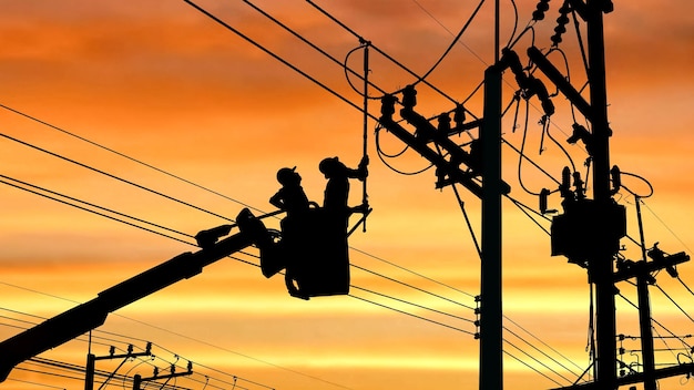 Silhouette two electricians on crane truck installing electric\
transmission on power pole at sunset