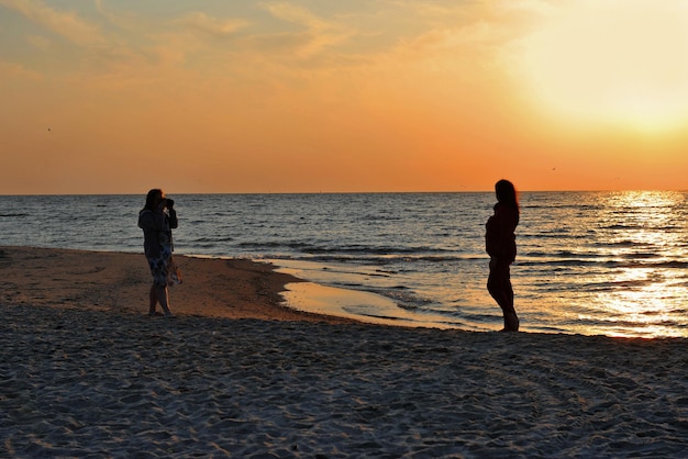 Silhouette of two girls taking pictures on the sea coast against the background of the rising mornin