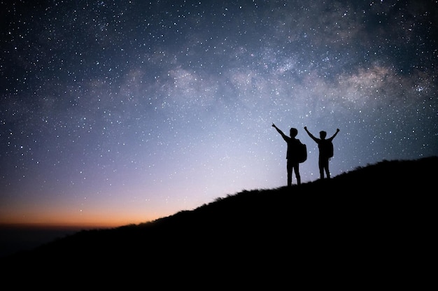 立って星の天の川と山頂の夜空を眺める2人の観光客のシルエット彼らは旅行を楽しんでおり、彼が頂上に到着したときに成功しました