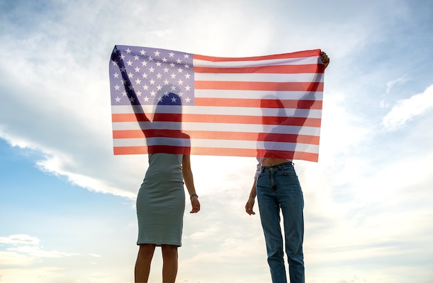 Silhouette of two young friends women holding USA national flag in their hands at sunset. Patriotic girls celebrating United States independence day. International day of democracy concept.