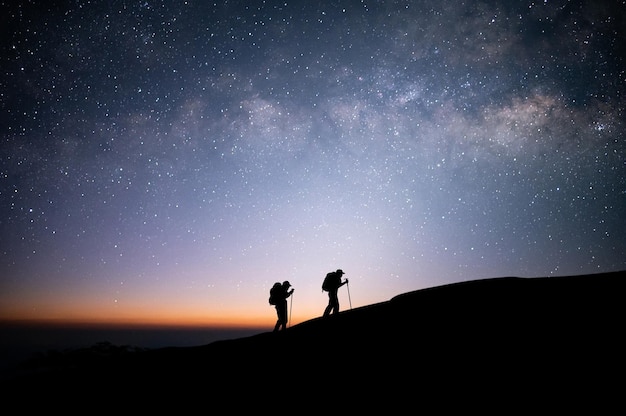 Silhouette of two young traveler and backpacker hiking to the\
top of the mountain with beautiful view star milky way over the sky\
he enjoyed traveling and was successful when he reached the\
summit