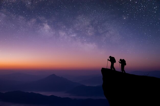 Foto silhouette di due giovani viaggiatori e backpacker in piedi sulla scogliera guardato la stella e la via lattea in cima alla montagna ci è piaciuto viaggiare e ha avuto successo quando ha raggiunto la vetta