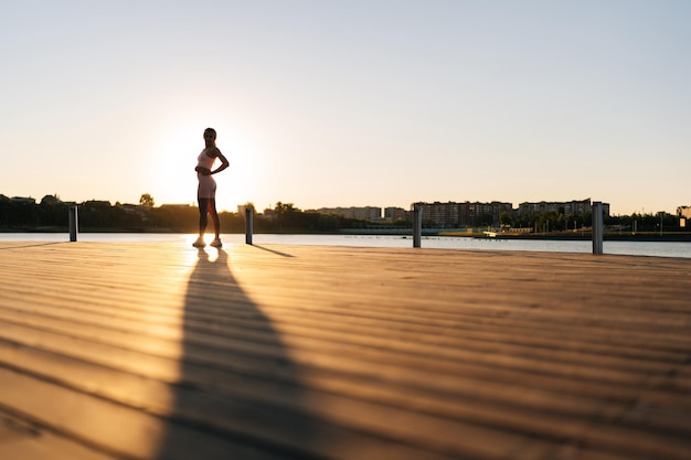 Silhouette of unrecognizable woman in sportswear warming up muscles practicing stretching exercises running or working out by water in city park