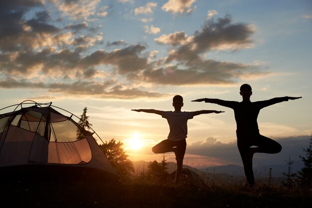 Silhouette of woman and boy standing in yoga pose at sunset near the camping