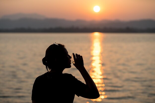 夕暮れの空に沿って海に立ってガラスからアルコールを飲む女性のシルエット