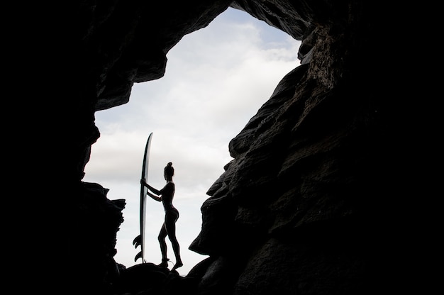 Silhouette donna ragazza in costume da bagno in piedi con il surf all'interno della roccia sulla spiaggia oceano atlantico