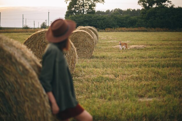 Silhouette of woman in hat looking at ginger dog at hay bale in summer evening in field