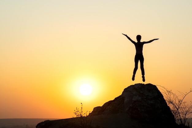 Silhouette of woman hiker jumping alone on an empty rock at sunset