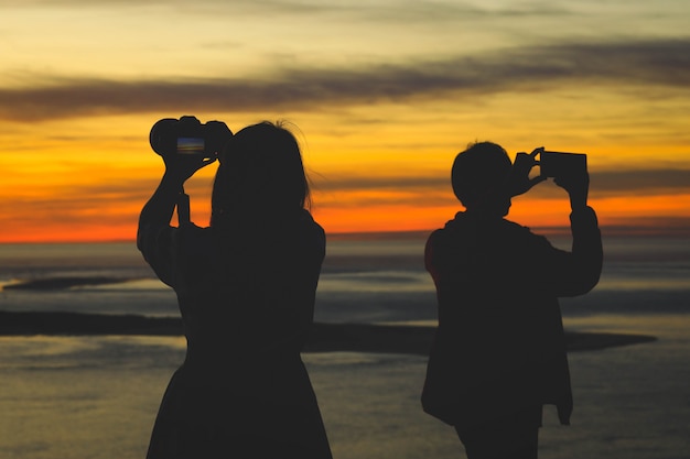 日の出または日没時に彼女の友人と写真を撮るカメラを保持している女性のシルエット
