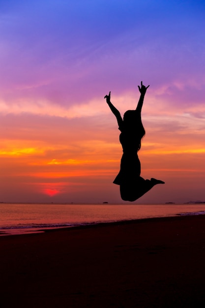 Siluetta della donna che salta con le mani in su sulla spiaggia del mare al tramonto