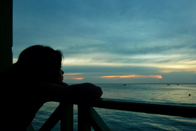 Silhouette woman leaning on bridge railing by sea against sky during sunset