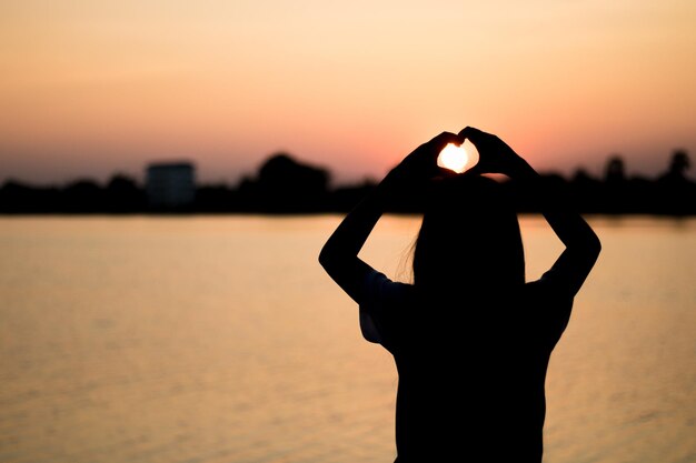 写真 夕暮れの空を背景に湖の近くで手で心の形を作る女性のシルエット