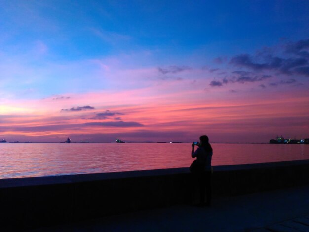 Silhouette woman photographing sea against sky during sunset