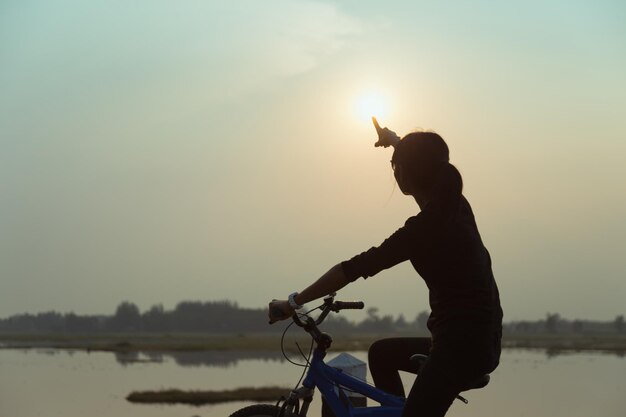 silhouette woman riding bicycle at lake against sky during sunset