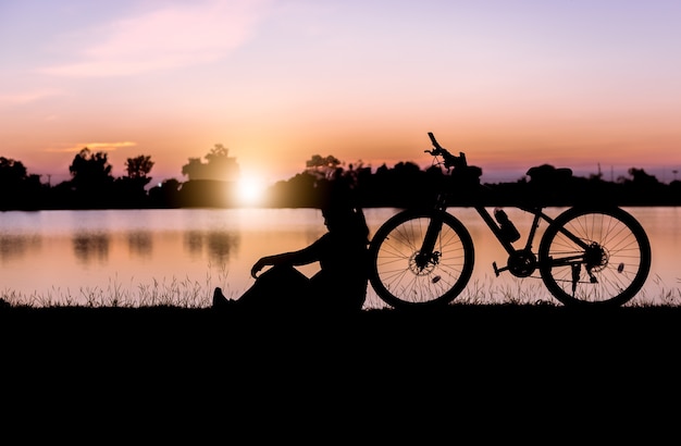 シルエットの女性は日没の自転車の近くに座っています。