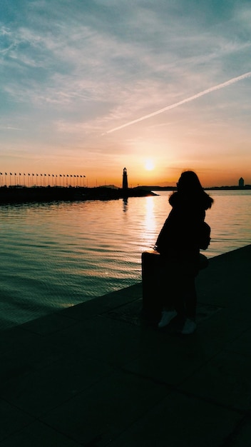 Silhouette woman sitting by sea against sky during sunset