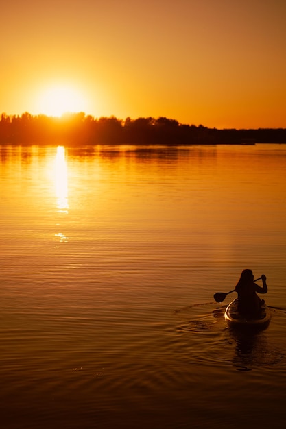 Silhouette of woman sitting on paddle board with oar with hands rowing straight on beautiful lake with astonishing sunset in background mirroring in water