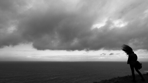 写真 雲の空を背景に海の崖の上に立っているシルエットの女性