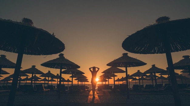 Silhouette of woman standing with head in hands by lounge chairs and parasols at beach during sunset