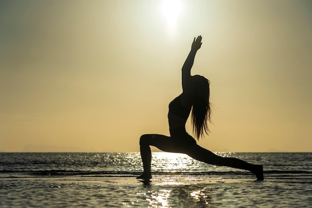 Силуэт женщины, стоящей в позе йоги на тропическом пляже во время заката Девушка практикует йогу у морской воды