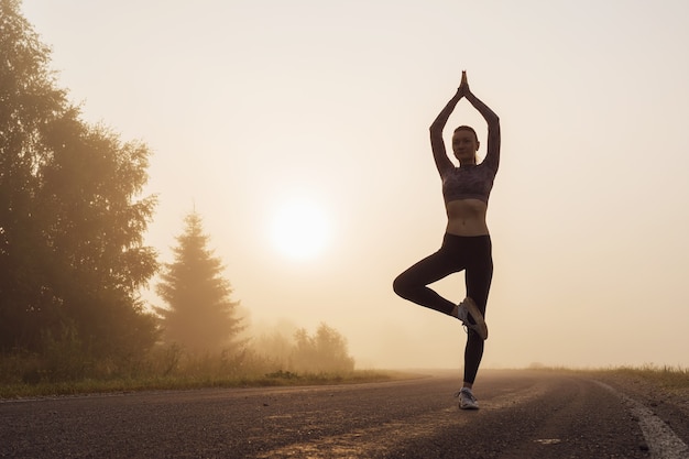 Silhouette of a woman at sunrise doing vrikshasana exercise near the edge of the road, tree pose