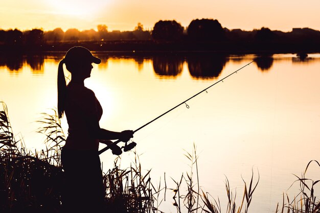 Silhouette of a woman at sunset with a fishing rod near the pond