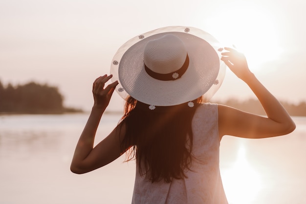 The silhouette of a woman at sunsetback view of a young woman holding the brim of a straw hat with h...