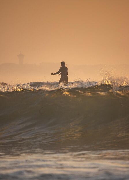 해가 지는 동안 하늘을 배경으로 바다에서 서핑하는 실루 여성